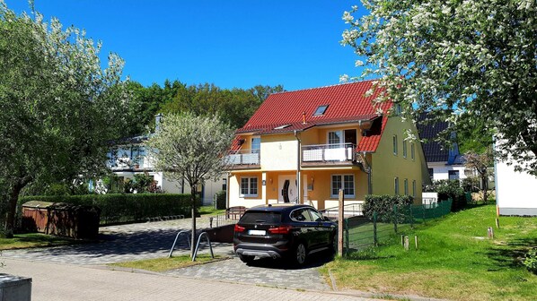 Une maison à deux étages avec un toit recouvert de tuiles rouges, un balcon et une voiture garée dans l’allée.