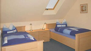 Quarto com duas camas de solteiro, uma escrivaninha, uma cadeira, um abajur e um claraboia.