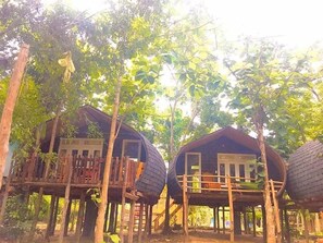 Dos cabañas de estilo casa árbol con barandillas de madera y techo de paja, rodeadas de follaje denso.