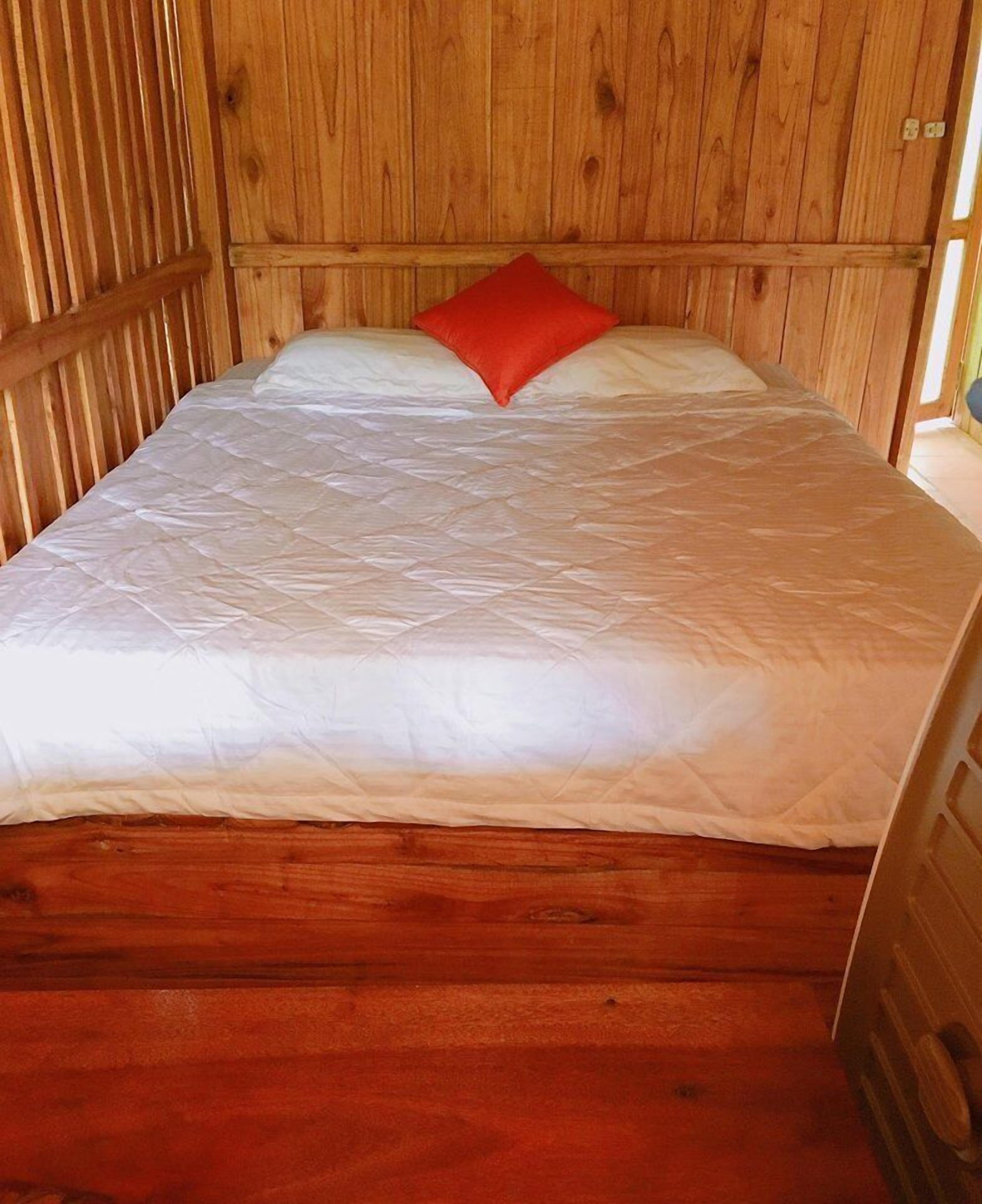 Una cama de madera con un colchón blanco y una almohada roja en una habitación con paredes de madera.