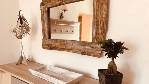A wooden-framed mirror on a wall, a decorative plant in a black pot, and a wooden tray on a wooden counter.