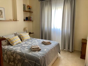 A neatly arranged bedroom with a bed, pillows, a bedside table, a lamp, and a shelf with books and a vase.