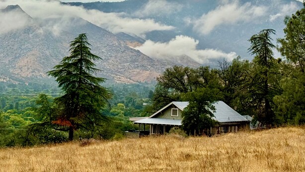 Una casa con techo inclinado, rodeada de hierba alta y árboles, con montañas al fondo.
