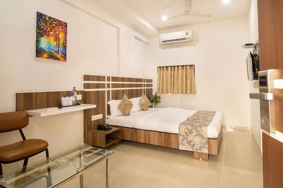 A hotel room with a bed, a desk, a chair, a glass table, a painting on the wall, and an air conditioning unit.