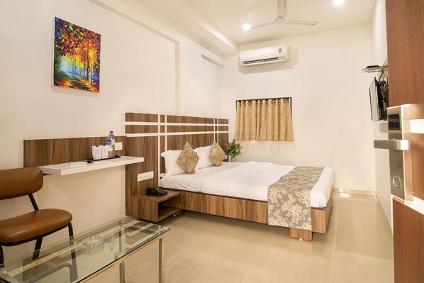 A hotel room with a bed, a desk, a chair, a glass table, a painting on the wall, and an air conditioning unit. - Hotel Sheetal Plaza (Surat)