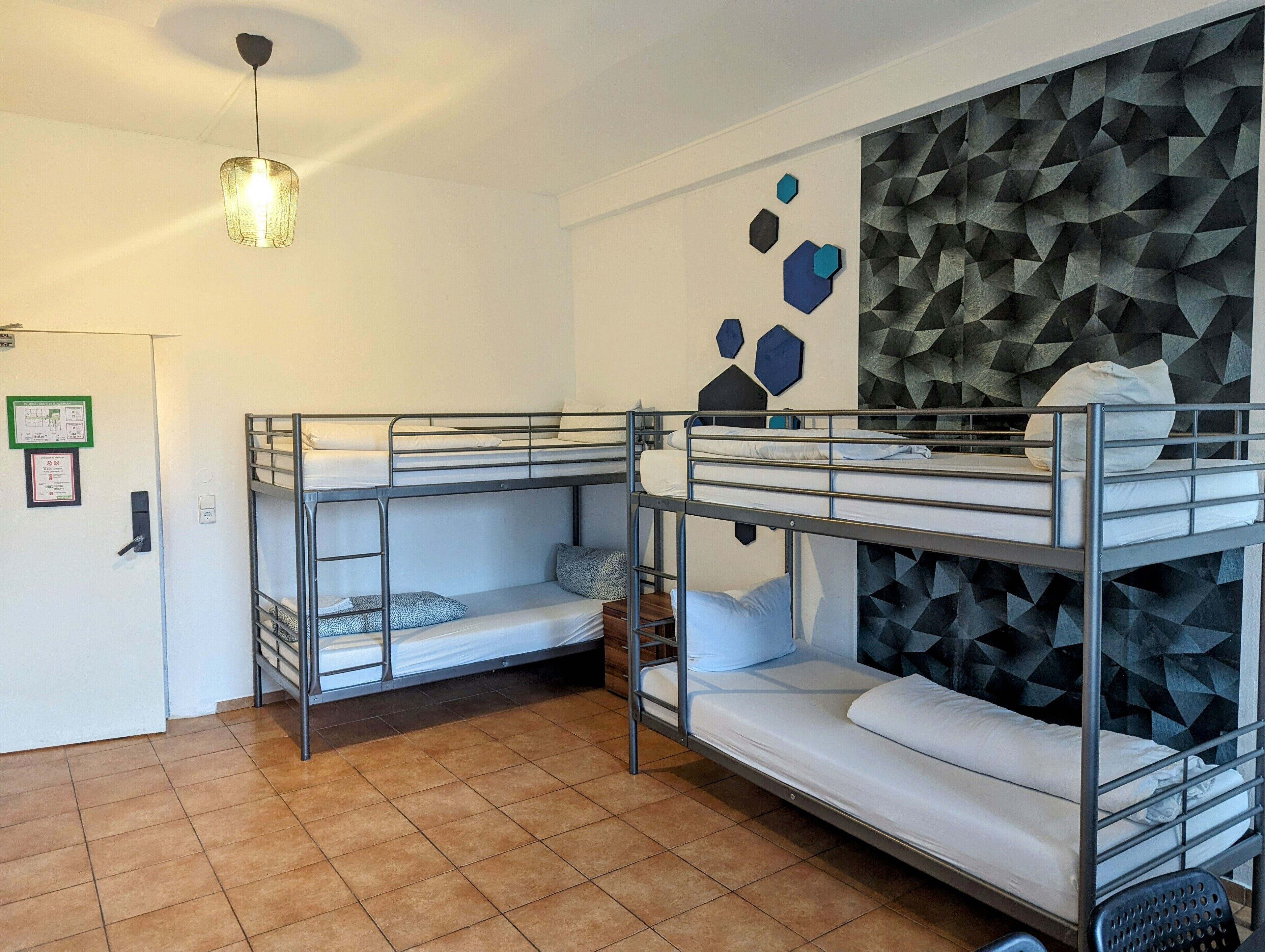 A dormitory room with bunk beds, a wall-mounted geometric pattern, and a hanging light fixture.
