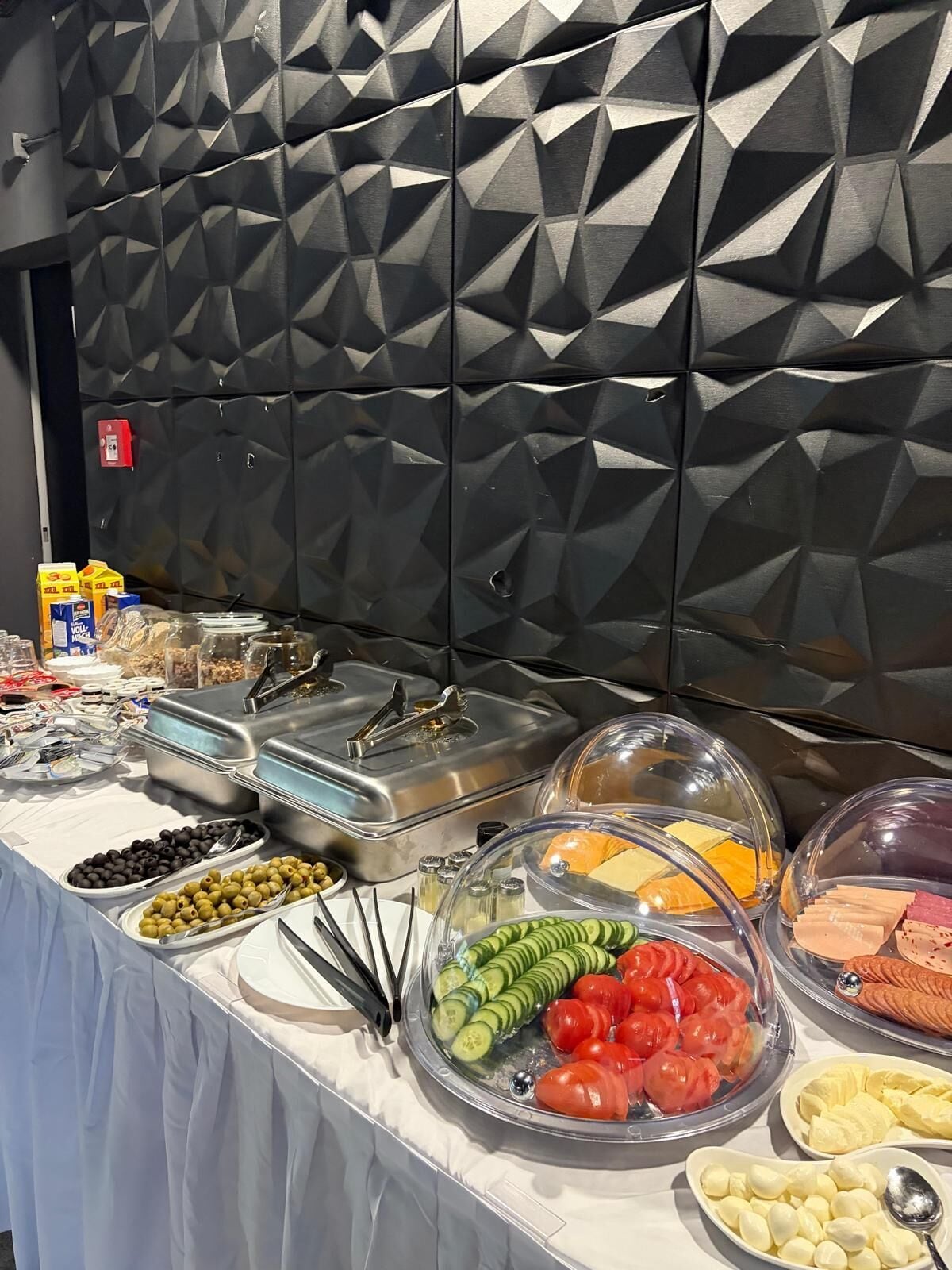 Un buffet proposant une variété de plats, notamment des olives, des concombres, des tomates et du fromage, avec un mur aux motifs géométriques en arrière-plan.