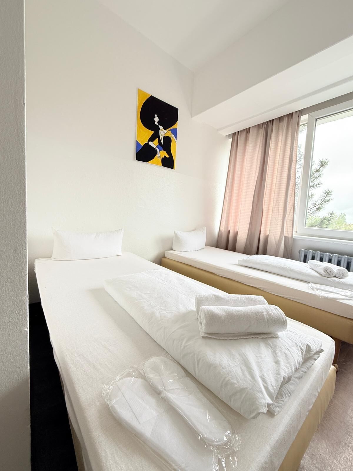 Una camera d'albergo con due letti, una finestra con tende, un termosifone e un quadro appeso al muro.
