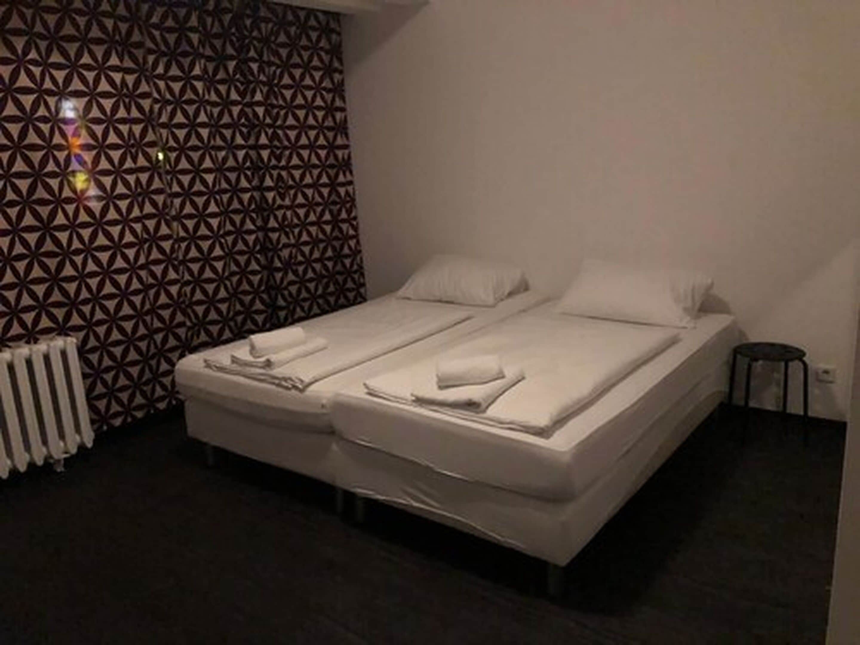 A hotel room with a patterned wall, a bed with white linens, a radiator, and a small stool.