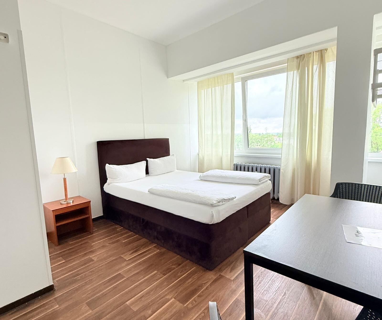 A hotel room with a bed, a table, a lamp, and a window with curtains.