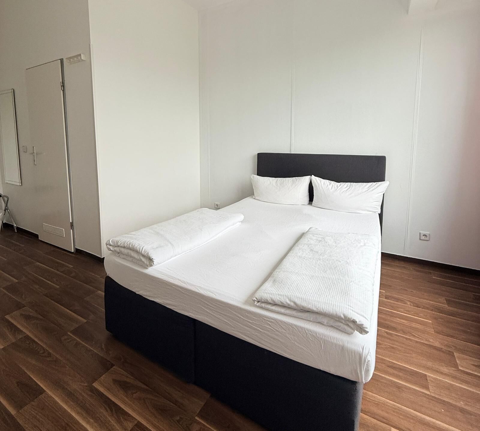 A neatly made bed with white linens, a dark headboard, and a wooden floor.