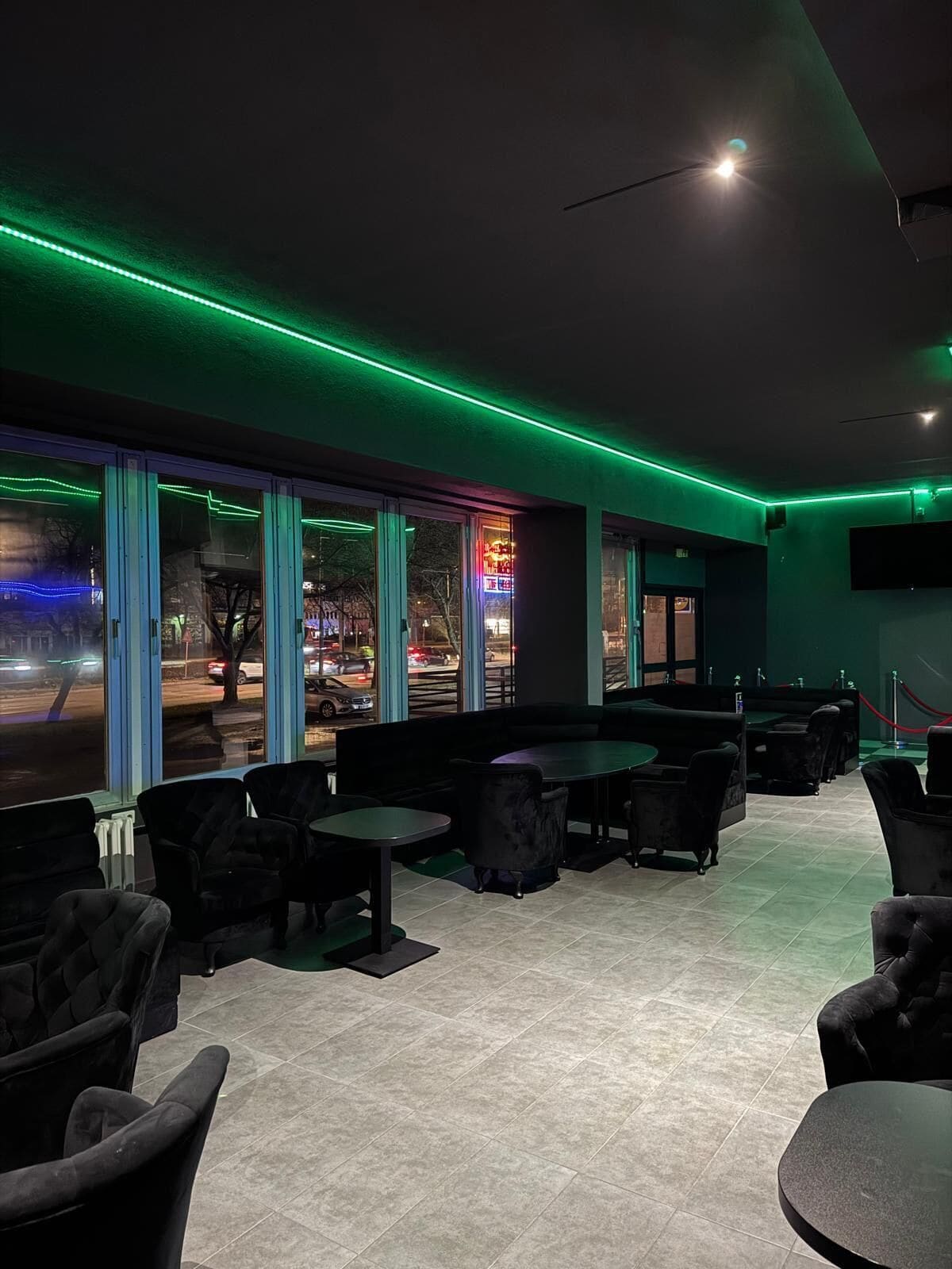 A dimly lit indoor space with large windows, green ambient lighting, and a variety of seating arrangements.