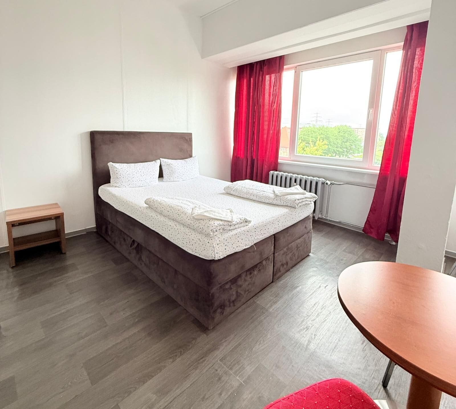 Un dormitorio con cama, una mesita, una ventana con cortinas y un radiador.