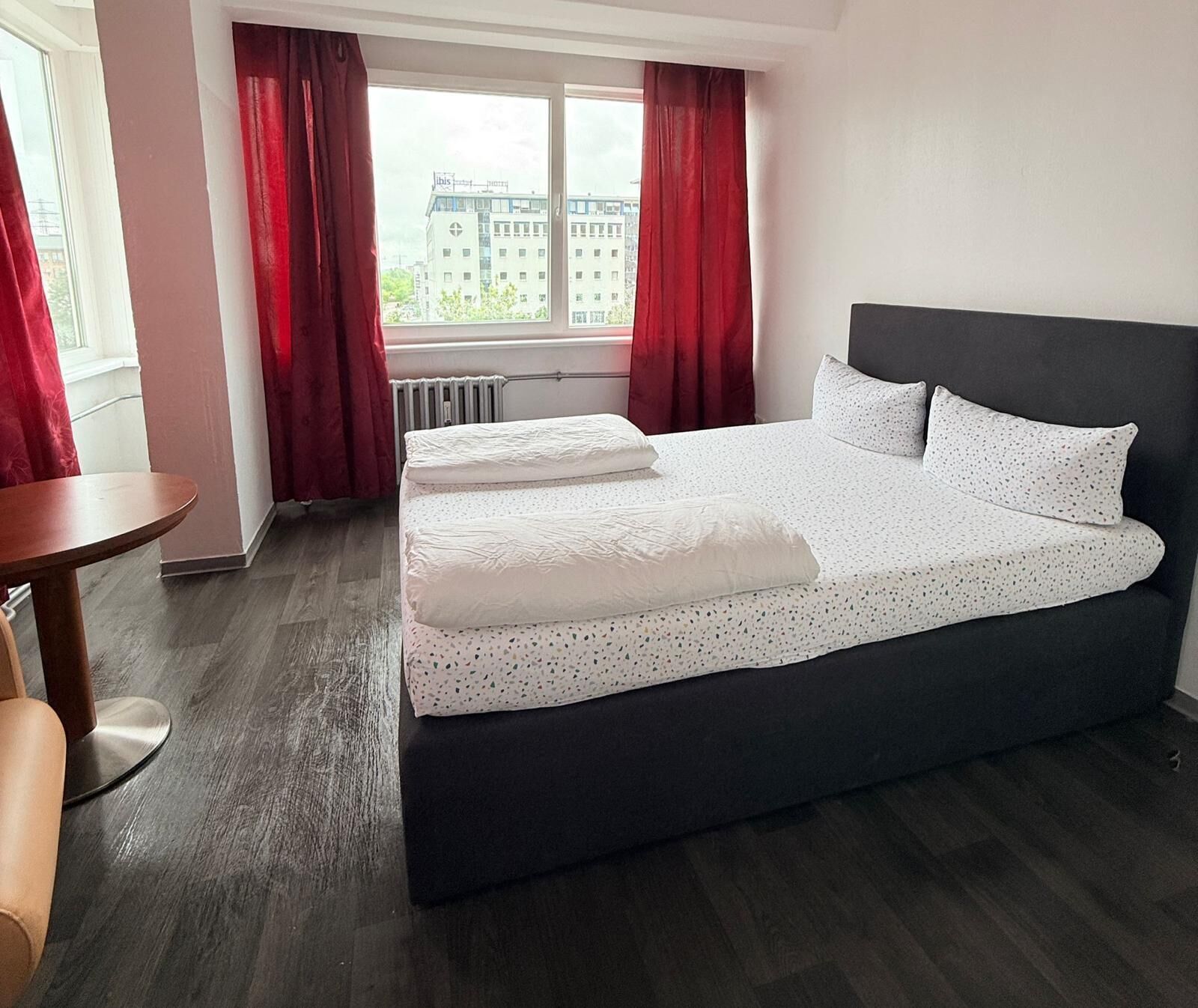 A hotel room with a large bed, a small round table, and a view of buildings through the window.