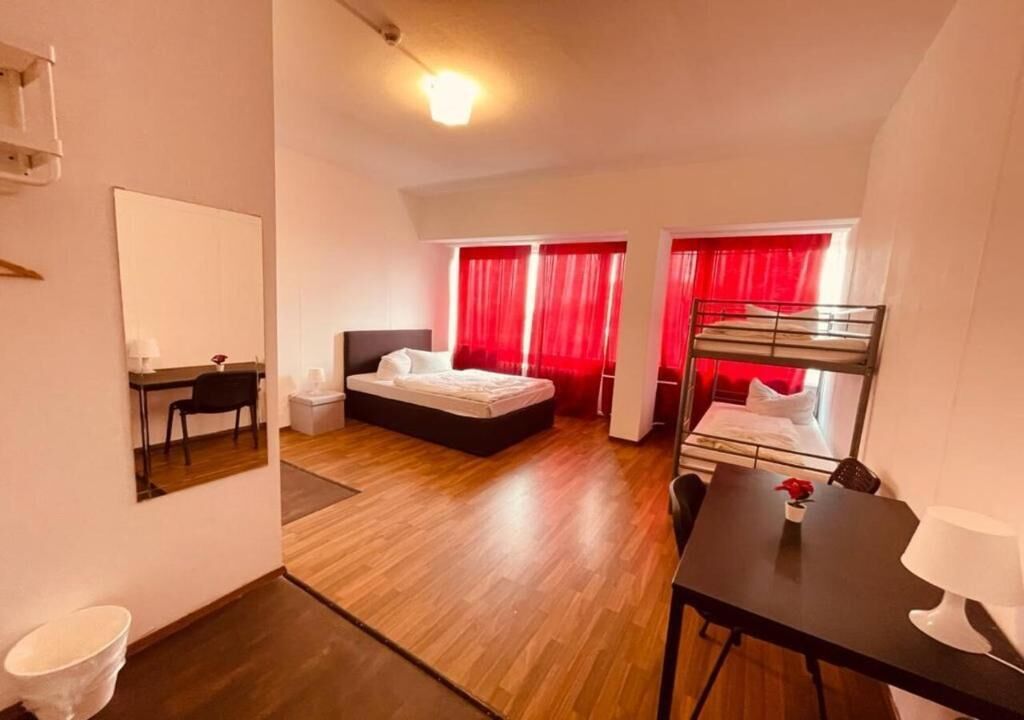 Habitación de hotel pequeña con literas, una cama individual, un escritorio con silla, una mesita con lámpara y un espejo.