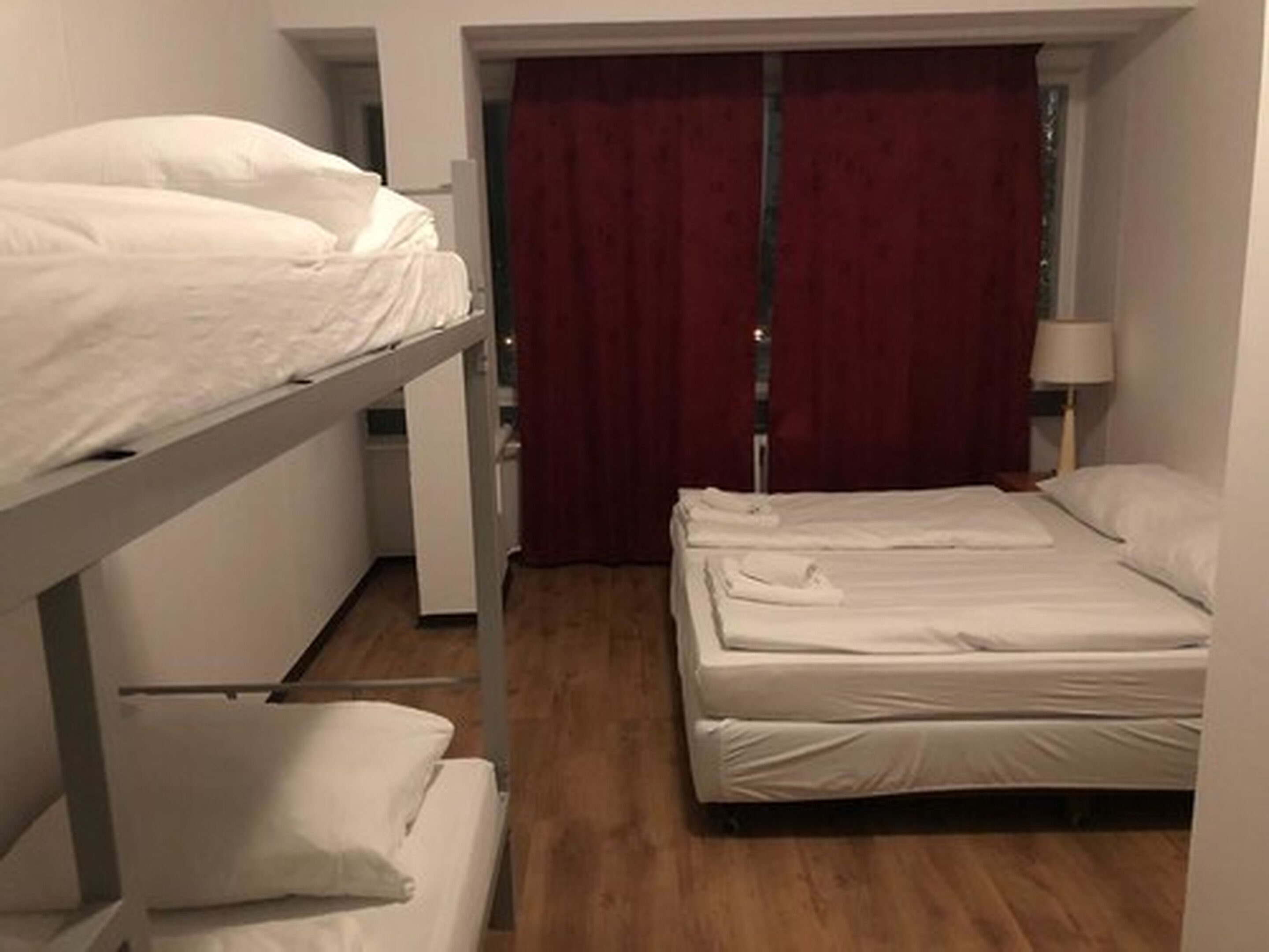 Una habitación con camas literas y una cama individual, suelo de madera, una cortina roja y una lámpara.