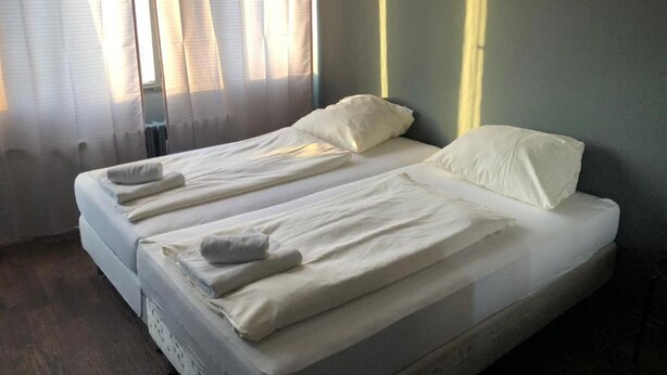 A single-bed room with a wooden floor, a large window with sheer curtains, and a plain wall. - Ootel.com (Berlin)