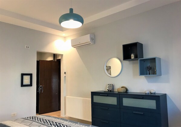 A bedroom with a bed, a dresser, a mirror, a wall-mounted air conditioner, and a door leading to another room.