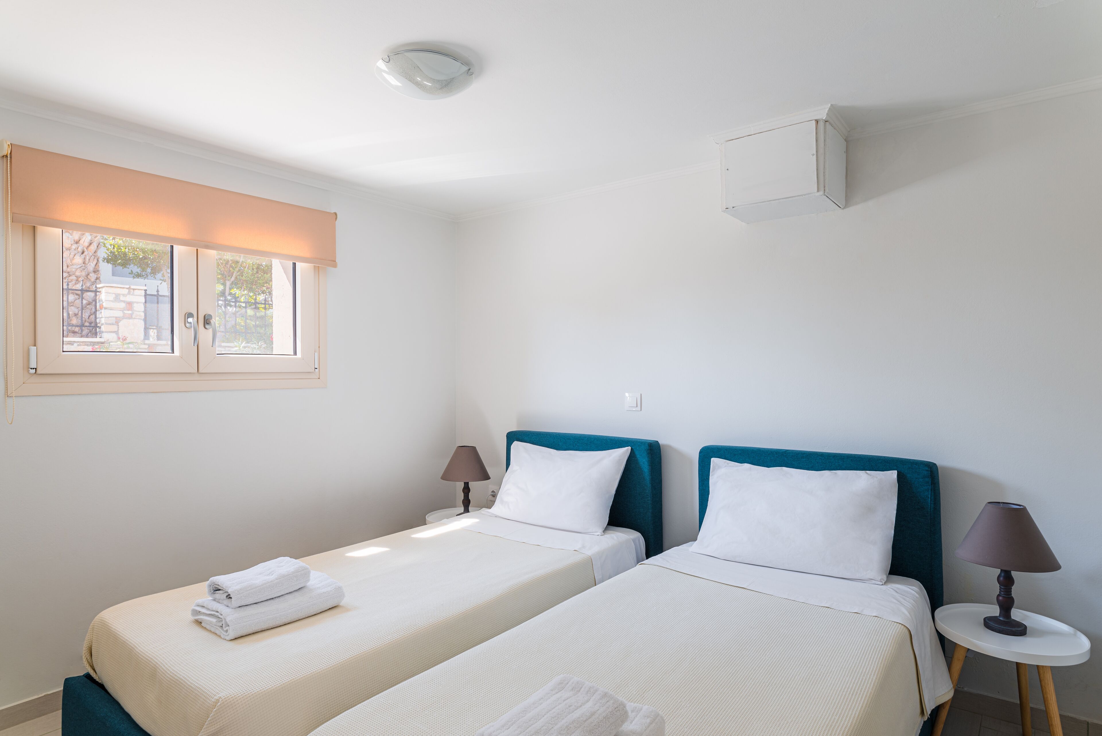 A hotel room with two beds, a window with a blind, a bedside table with a lamp, and a wall-mounted air conditioning unit.