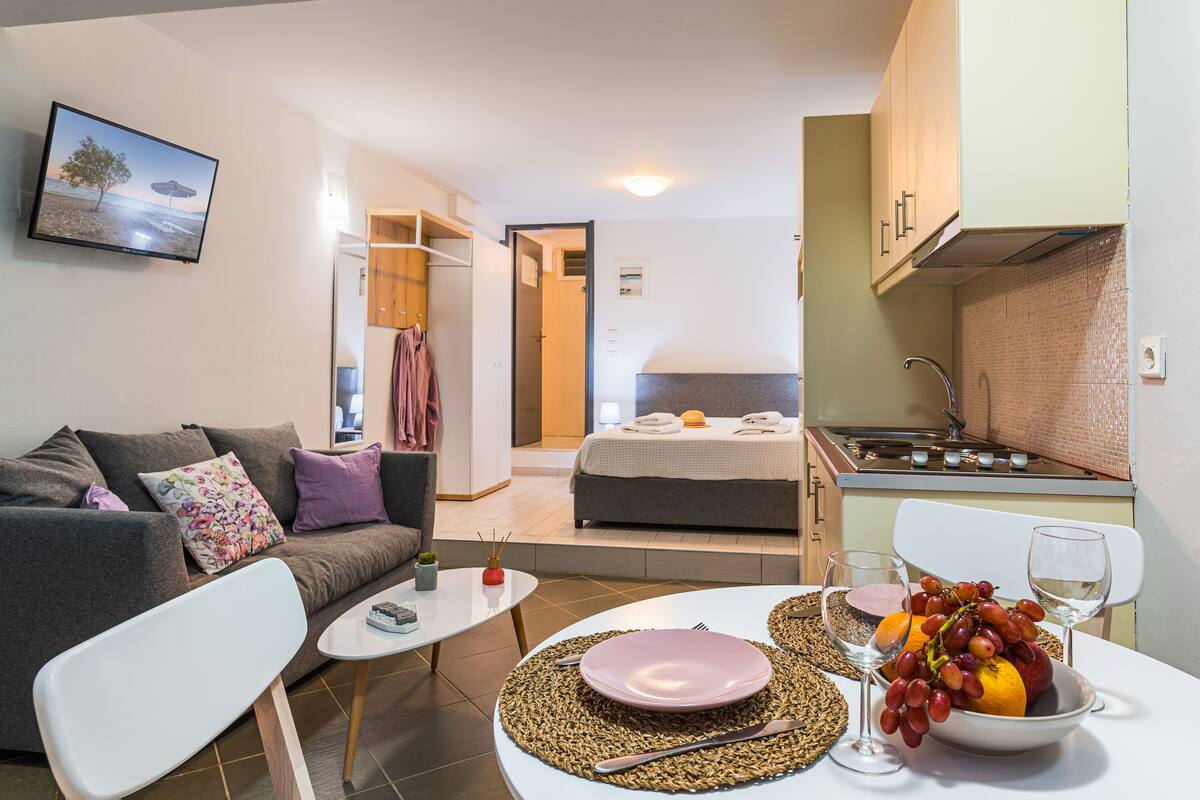 Un espacio habitable compacto con sofá, una pequeña mesa de comedor, una zona de cocina y un dormitorio con cama, y un baño visible al fondo.