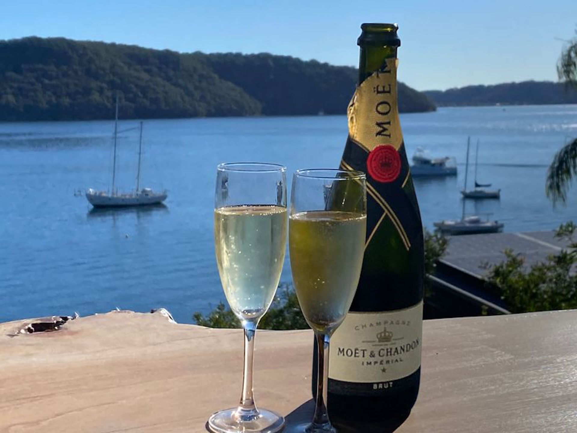 A bottle of Moët & Chandon champagne and two glasses on a table with a view of a calm body of water and boats in the background.