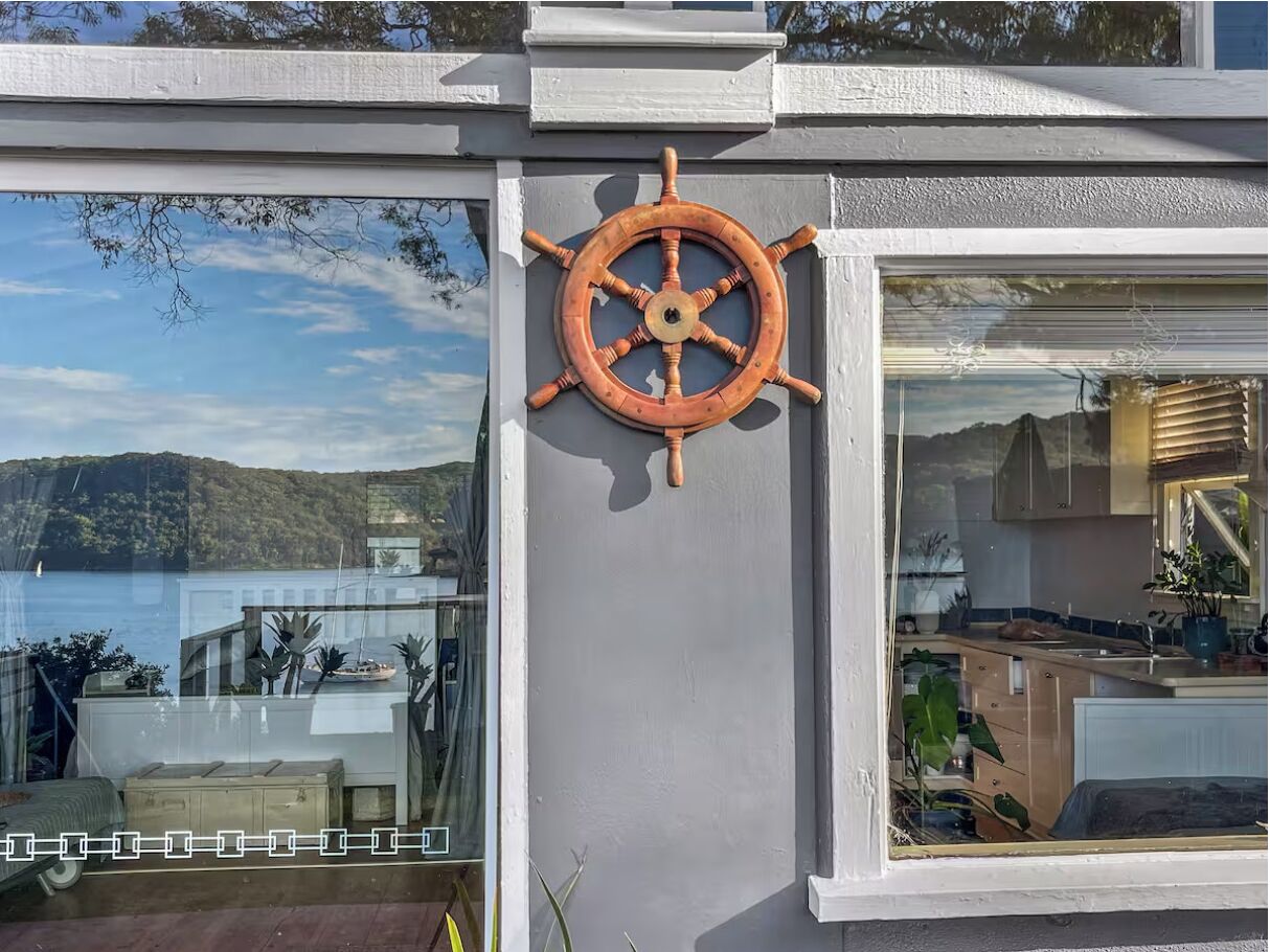 Une décoration murale sur le thème de la navigation, avec un gouvernail de navire, une porte vitrée donnant sur la mer et un aperçu d’un coin cuisine.