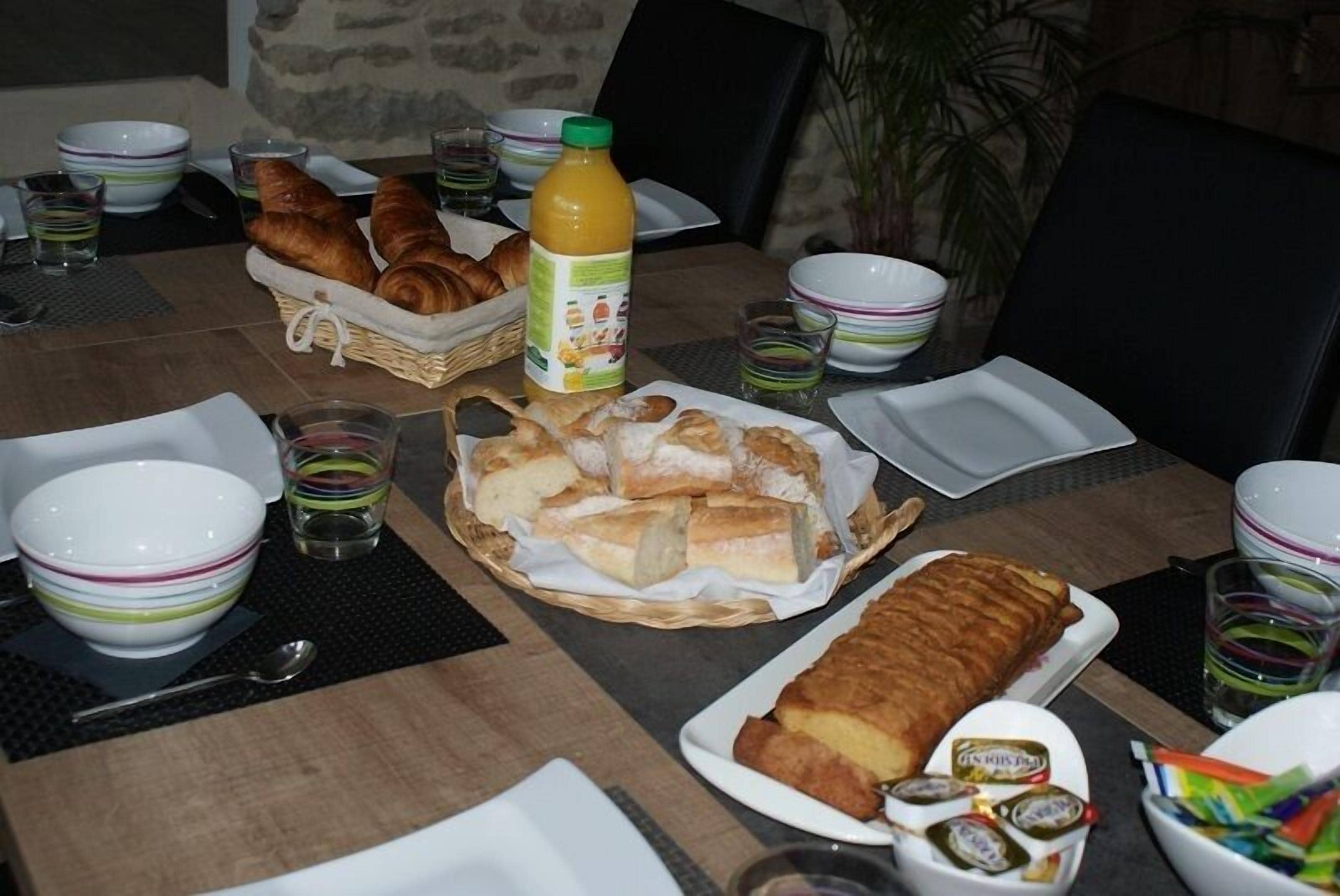 Una mesa de comedor servida con cruasanes, pan, una botella de jugo de naranja y diferentes fuentes y vasos.