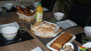 Una mesa de comedor servida con cruasanes, pan, una botella de jugo de naranja y diferentes fuentes y vasos.