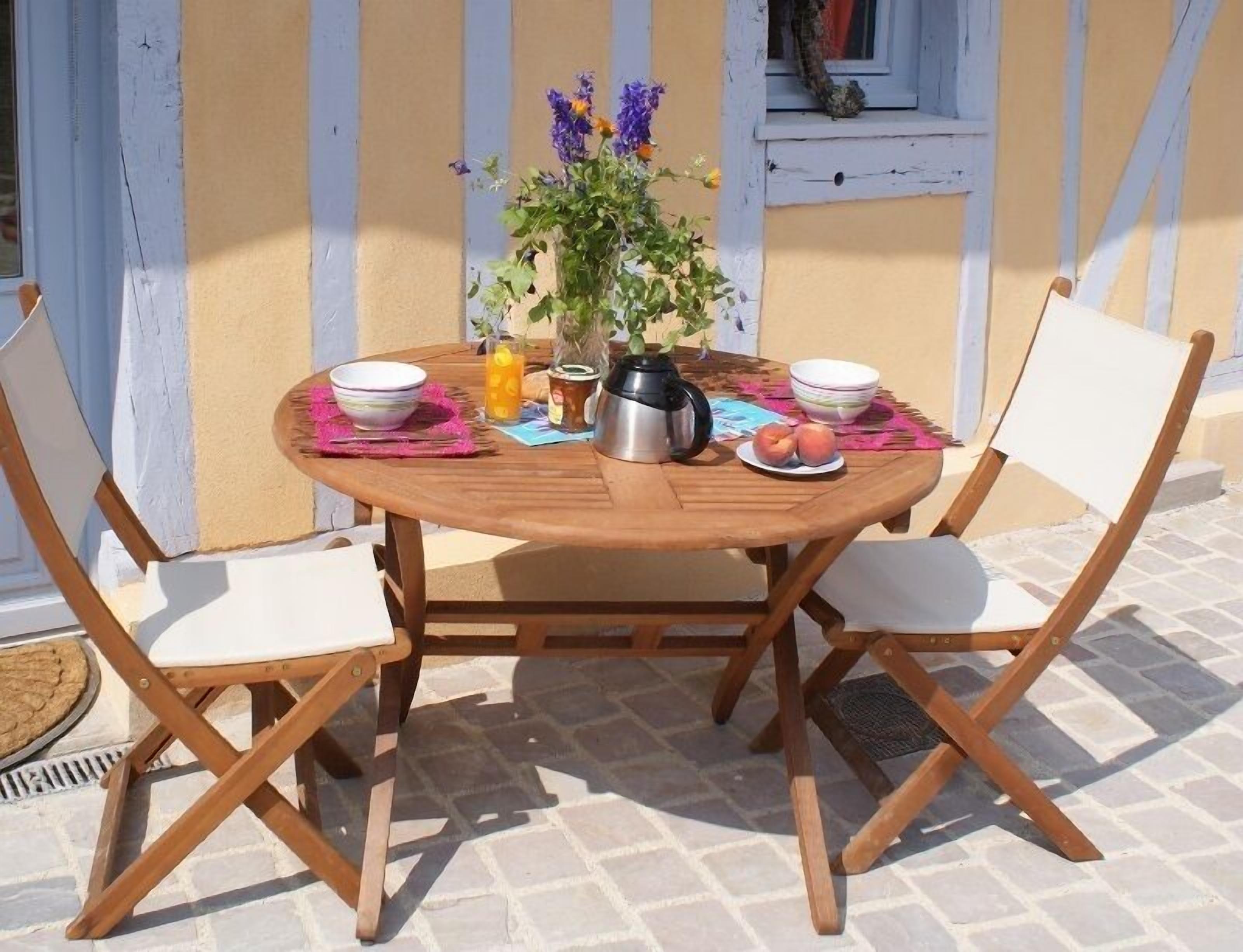 A round wooden table with two white cushioned chairs, a vase with flowers, a pitcher, and a plate with fruit.