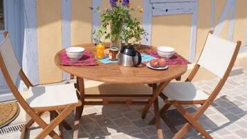 A round wooden table with two white cushioned chairs, a vase with flowers, a pitcher, and a plate with fruit.