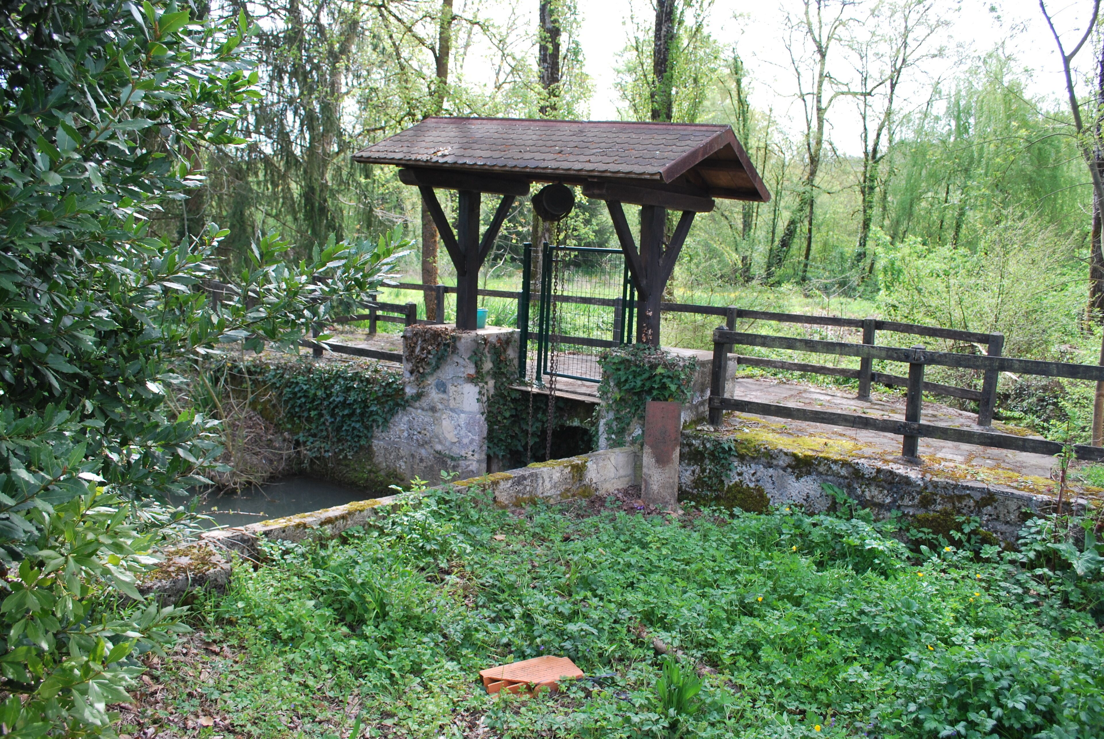Un belvédère rustique en bois, surmonté d’un toit en métal, entouré d’une végétation luxuriante et bordé d’un petit ruisseau.