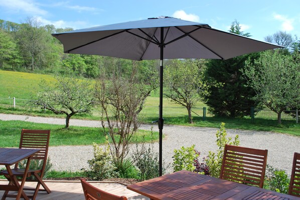 Eine Außenpatio mit Tisch und Stühlen, einem großen Sonnenschirm und Blick auf eine grüne Landschaft mit Bäumen und einem Zaun.