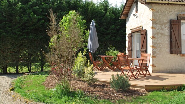 Una casa rústica con una terraza de madera, una pequeña mesa y sillas, y un área de estar al aire libre techada.