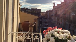 Ein Balkon mit schmiedeeisernem Geländer, Blumentöpfen und Blick auf eine Straße mit Gebäuden und Autos.