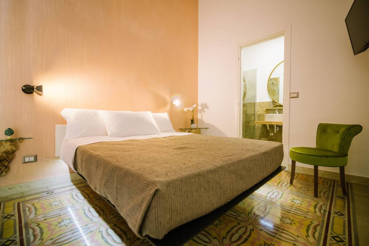 Ein ordentlich bezogenes Bett mit einer beigefarbenen Tagesdecke, ein grüner Sessel und ein Badezimmer mit Spiegel und Waschbecken.