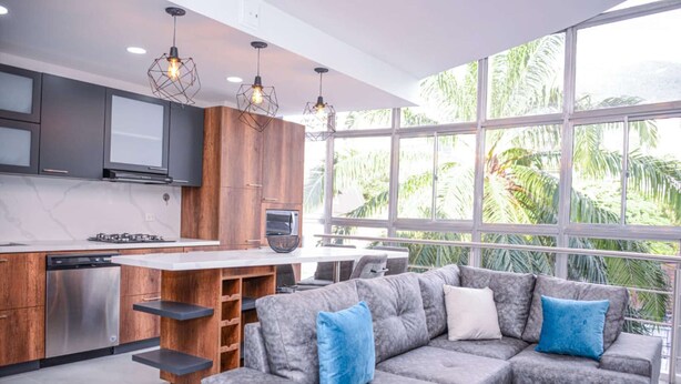 A modern kitchen with wooden cabinets, a built-in oven, and a dining area with a grey sofa and blue cushions. - 3 BEDROOM PENTHOUSE MIDDLE OF PARK LLERAS PRIVATE ROOF TOP WITH HOT TUB AC (Medellin)