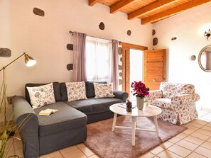 A cozy living room with a grey sectional sofa, a round coffee table, and a floral-patterned armchair. A wooden door and a mirror are visible in the background.