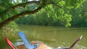 A wooden dock with two chairs and a table, overlooking a calm river surrounded by lush greenery.