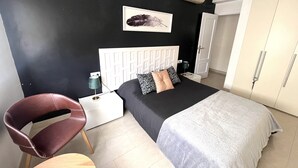 A modern bedroom with a black and white color scheme, a bed with a grey bedspread, a brown leather chair, a white bedside table, and a wall-mounted air conditioner. - Beautiful penthouse in the heart of Alicante (Alacant)