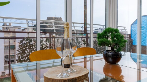 A glass table with a bottle of champagne, two wine glasses, and a bonsai tree on a balcony with a city view. - Beautiful penthouse in the heart of Alicante (Alacant)