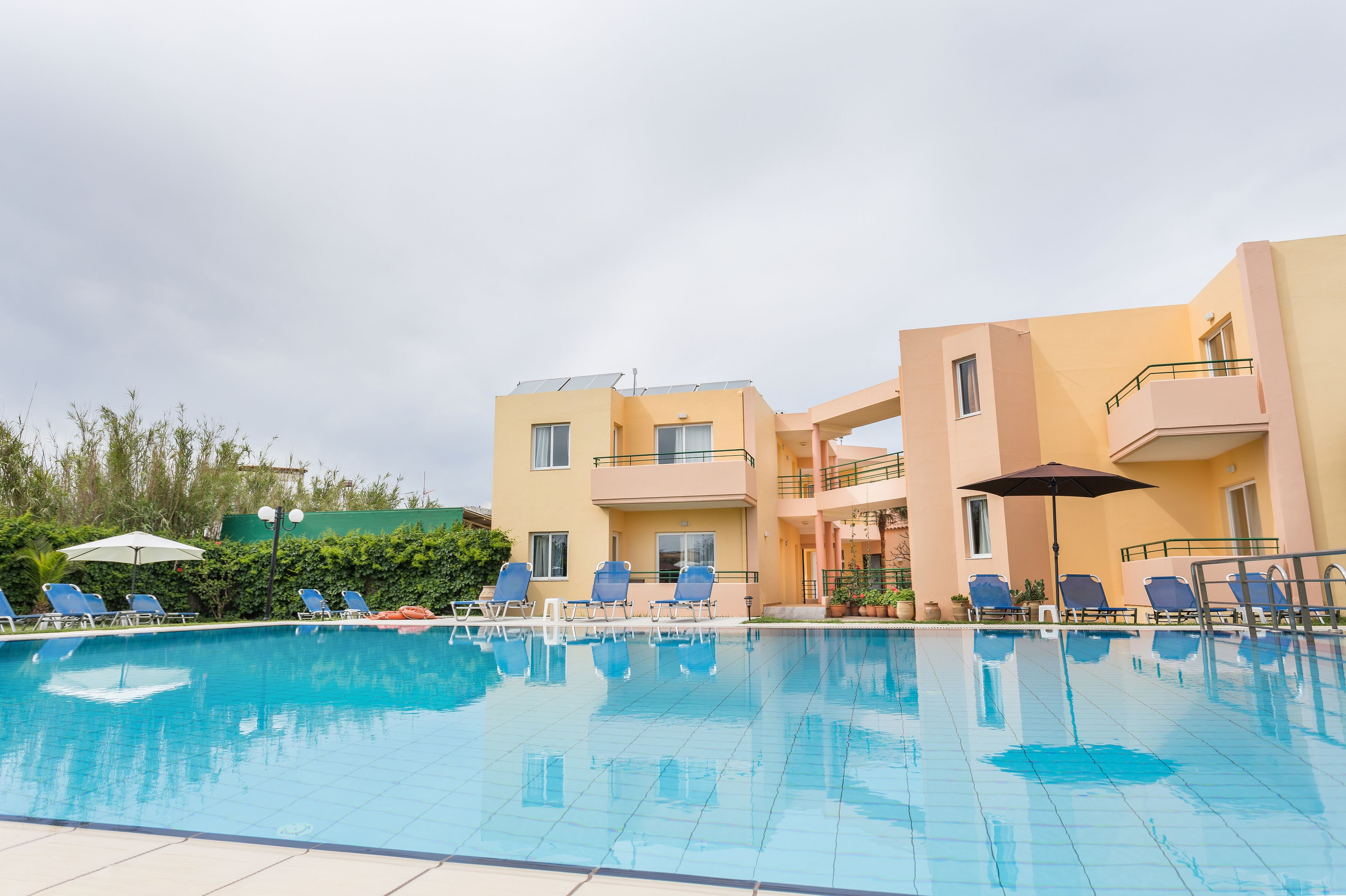 Une piscine entourée de chaises longues et de parasols, le tout bordé d’un bâtiment doté de balcons et d’une haie verdoyante.