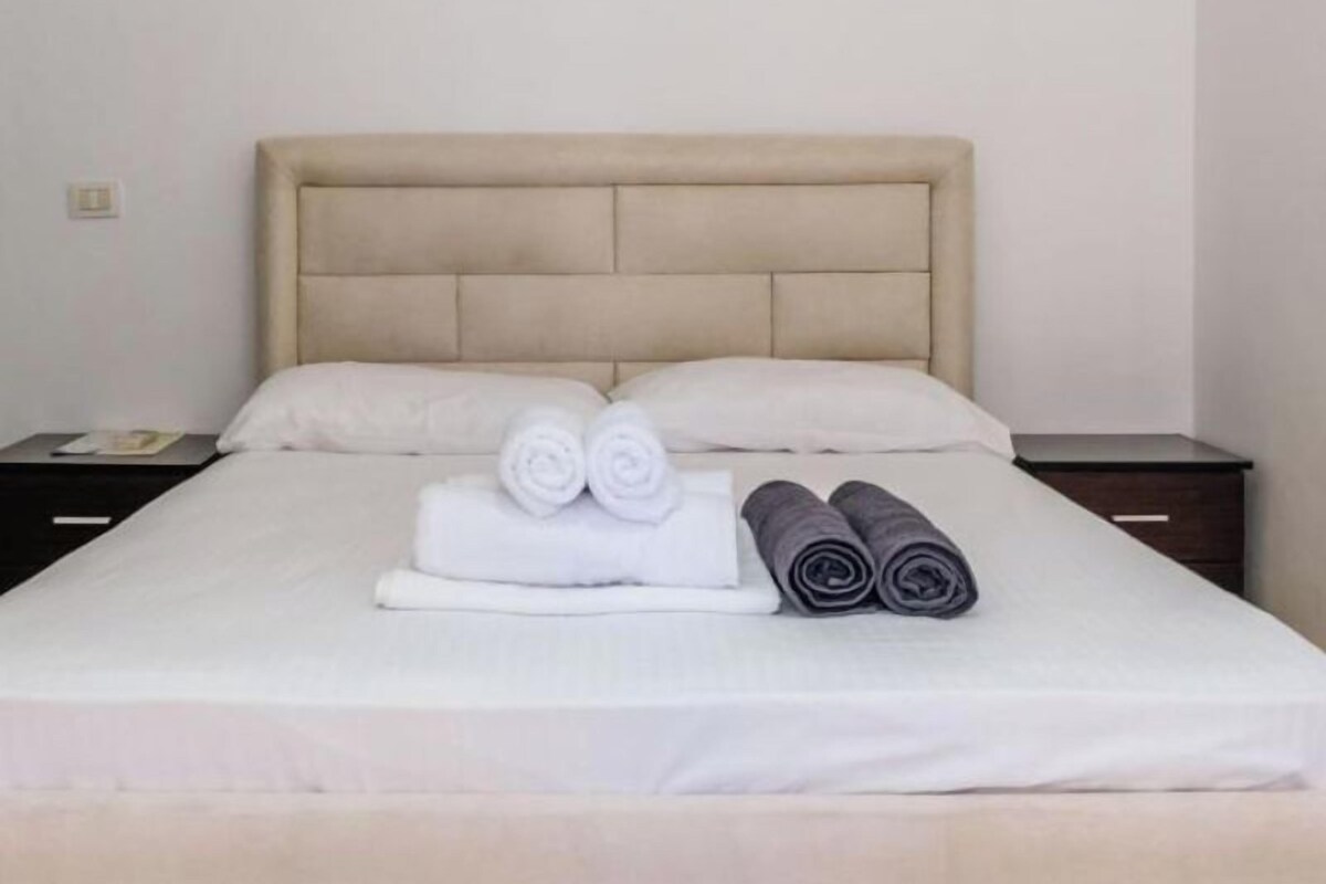 A neatly made bed with a beige headboard and white linens, accompanied by rolled towels and a small bedside table with a lamp.