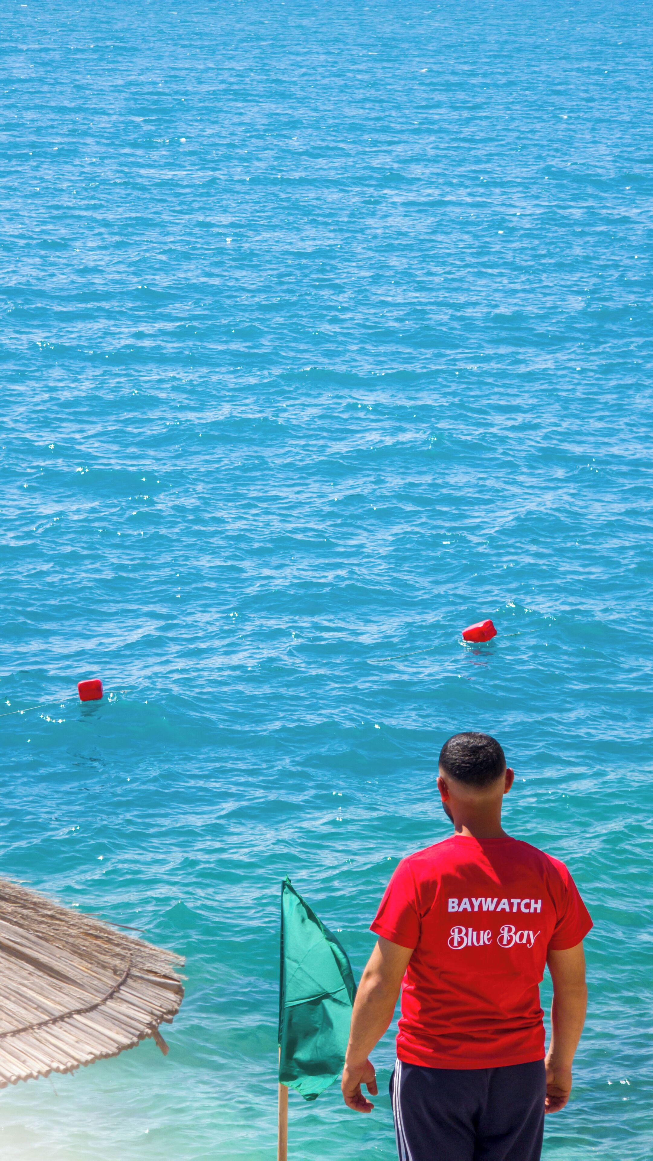 Una persona con camisa roja que lleva la inscripción "Baywatch Blue Bay" en la espalda, de pie junto a una masa de agua con una bandera verde en un asta.