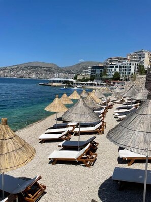 A beach with rows of sun loungers and thatched umbrellas, a clear blue sea, and a cityscape in the background. - Blue Bay Hotel (Sarandë)