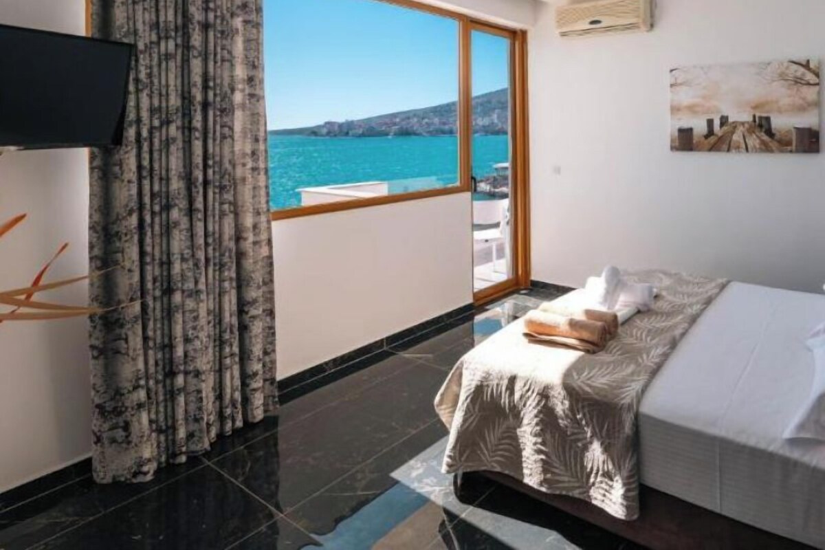 A hotel room with a bed, a TV, a large window with a view of the sea, and a painting on the wall.