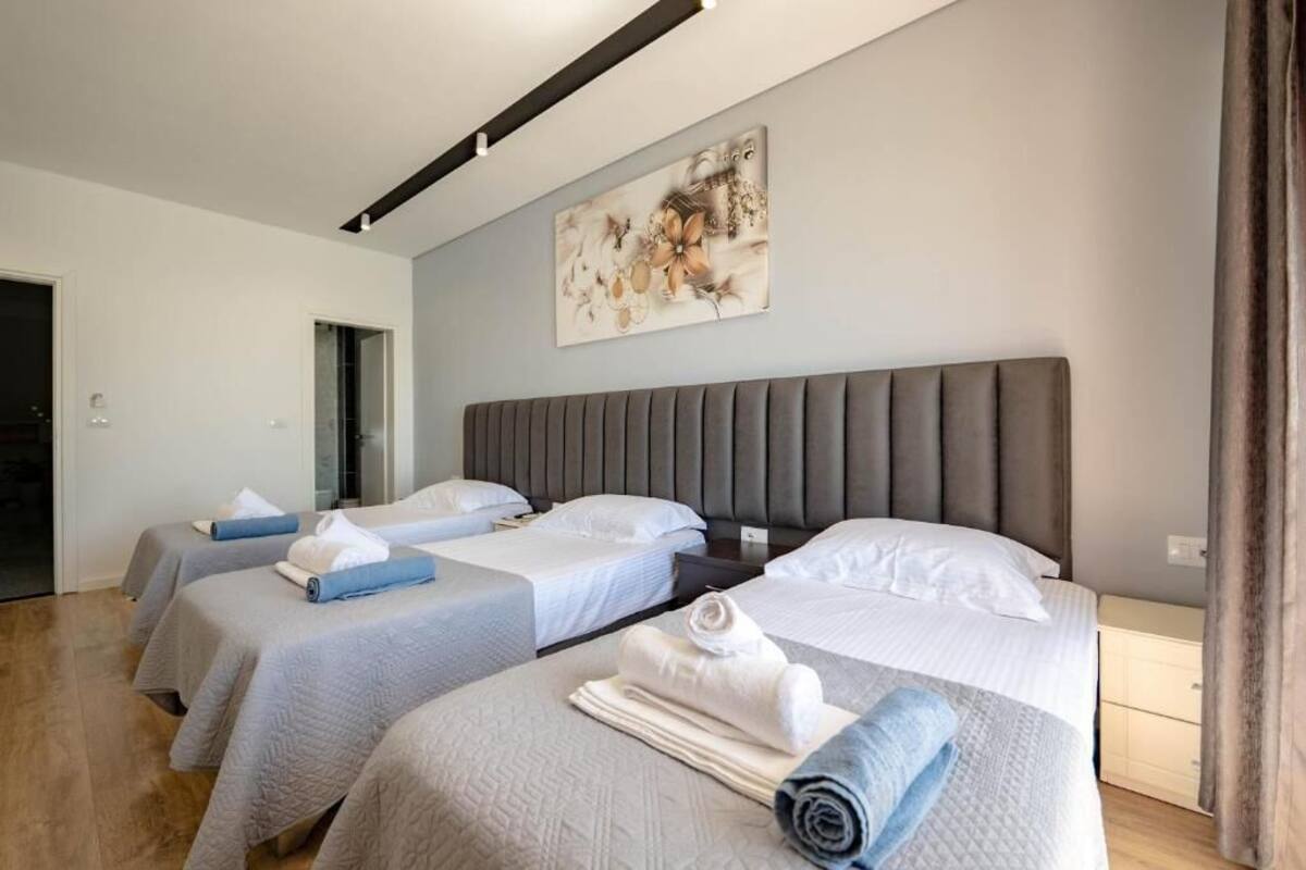 Ein Hotelzimmer mit drei Betten, jedes mit grauem Kopfteil und weißer Bettwäsche. Darüber an der Wand hängt ein Gemälde.