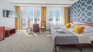 Habitación de hotel con cama, escritorio, silla, televisor y ventanas amplias con cortinas.