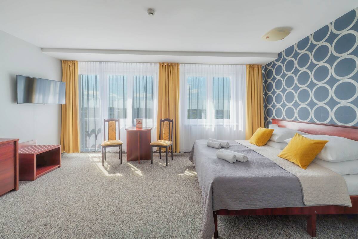 Hotelkamer met een bed, een bureau, een stoel, een televisie en grote ramen met gordijnen.