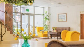 Un vestíbulo de hotel con una escalera, sillas amarillas, un sofá, una mesa con un florero y una planta colgante.