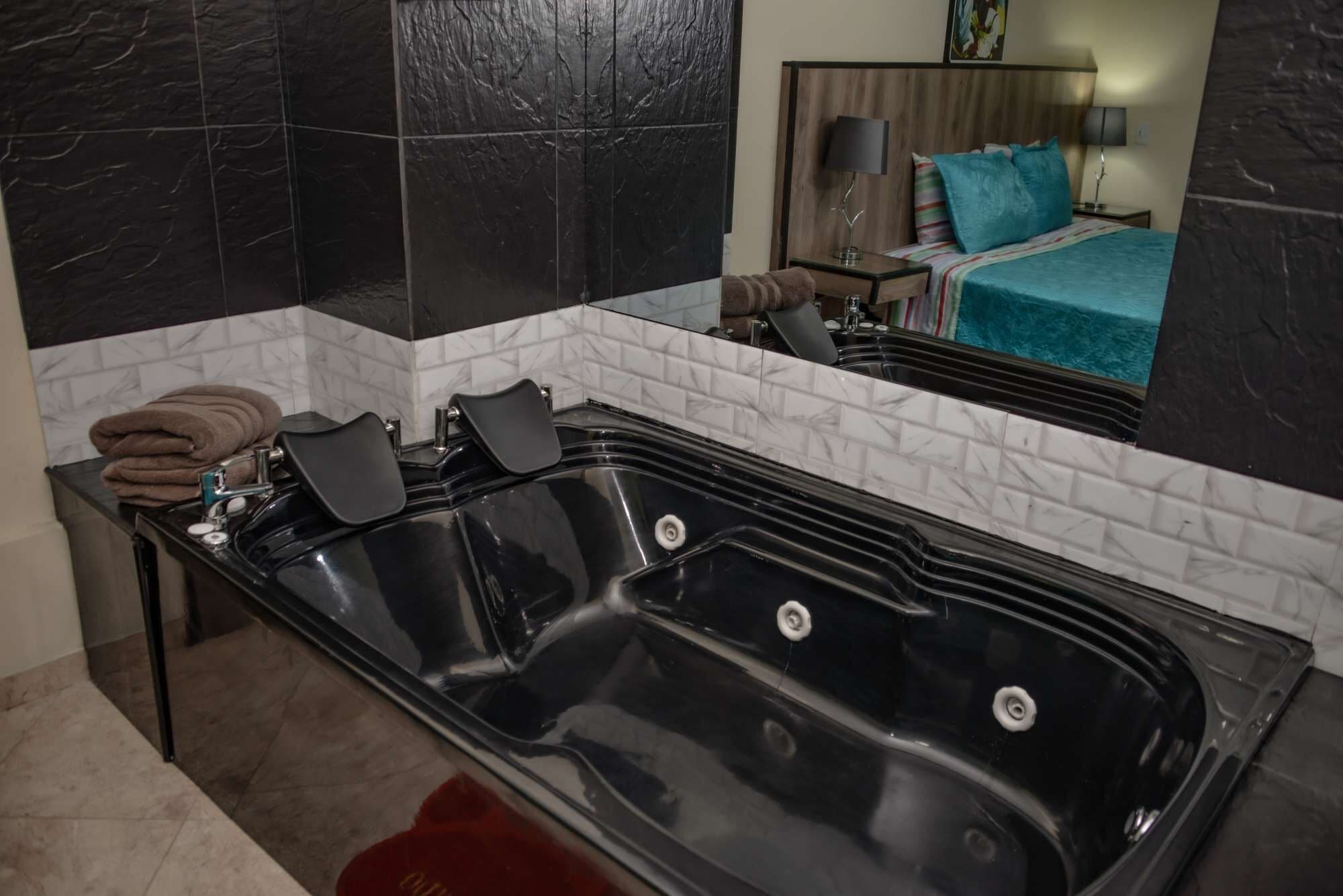 Ein modernes Badezimmer mit einer großen, schwarzen Badewanne, weißen Marmorfliesen und einem Spiegel, der ein Schlafzimmer mit einem Bett und einem Nachttisch reflektiert.
